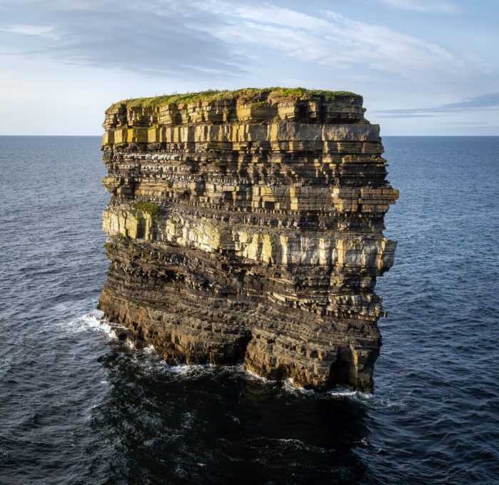 A towering rock formation rising sharply from the ocean, featuring layered cliffs with a green top and surrounded by water under a partly cloudy sky.